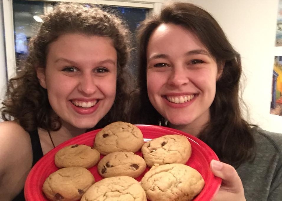 A photo of two smiling friends holding a plate of chocolate chip cookies.